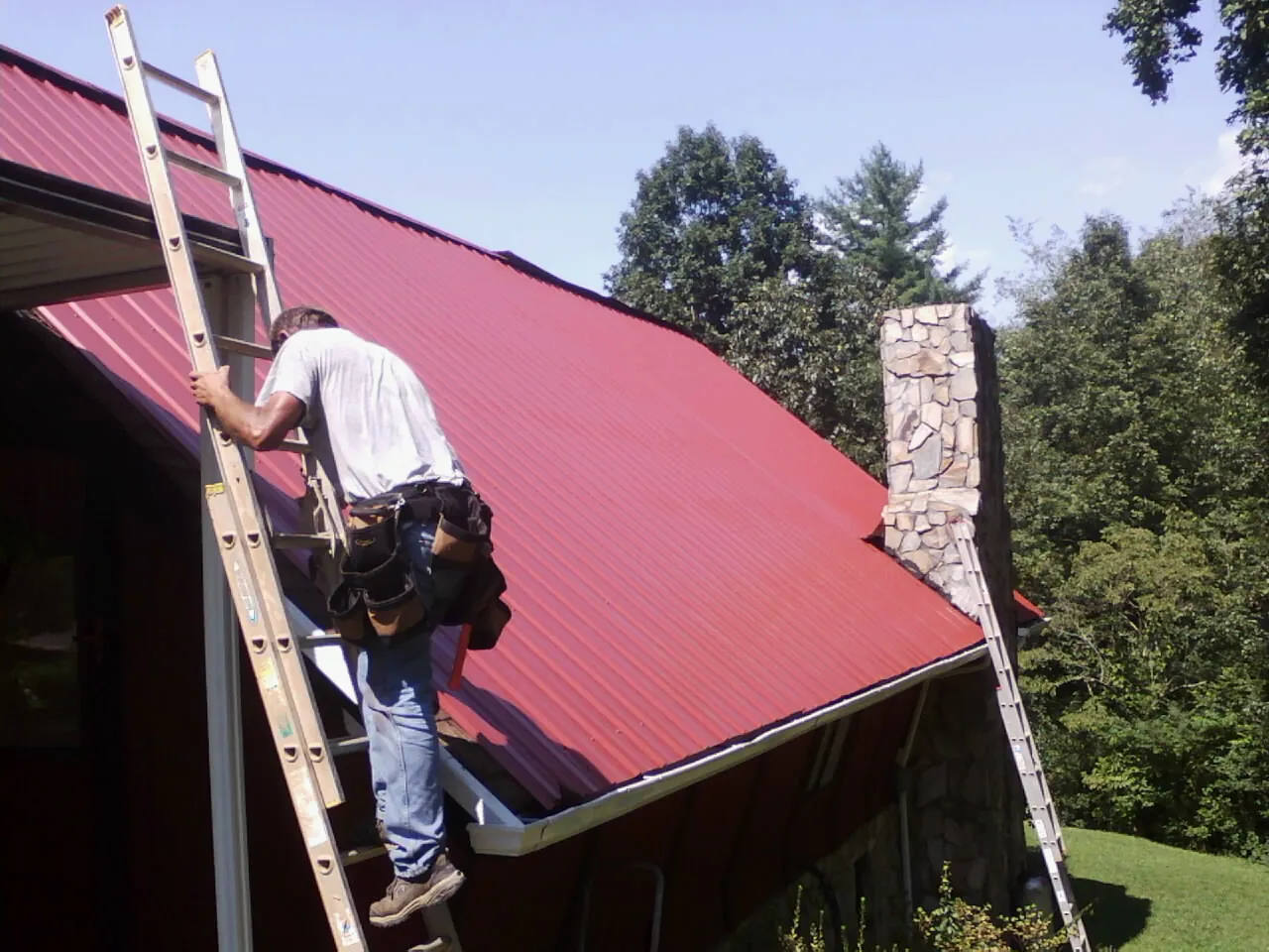 Roofer on a ladder beside a red metal roof and stone chimney — Soffit & Fascia Repair by Roofing Brothers in Fox Island